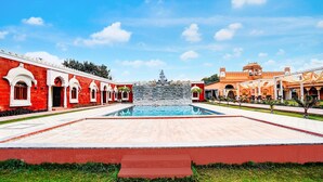 A pool area with a statue in the center, surrounded by red and orange buildings with arched windows. - Deluxehotel Banaras Kila (Pindra)