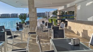 Un espace extérieur ensoleillé, aménagé avec des tables et des chaises, offrant une vue sur une plage et l’océan.