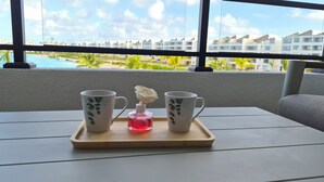 Un plateau avec deux tasses blanches ornées de motifs de feuilles vertes, une petite bouteille contenant un liquide rouge et un distributeur blanc en forme de rose, le tout posé sur une table en bois près d’une fenêtre donnant sur un complexe hôtelier.