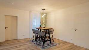Une pièce avec un plancher en bois, une table entourée de chaises, une fenêtre avec des rideaux et une porte.