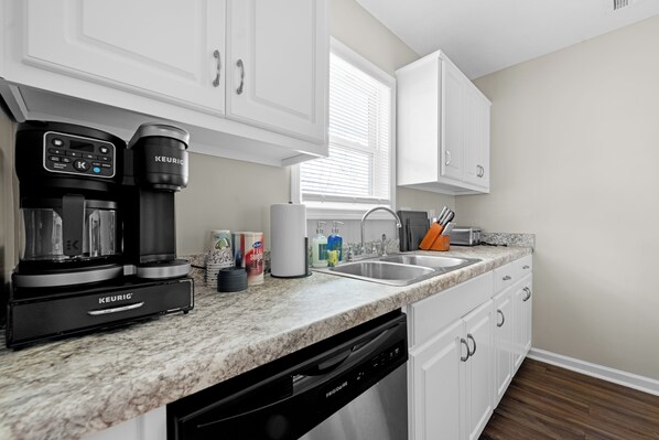 A kitchen with white cabinets, a coffee maker, a dishwasher, and a window with blinds.