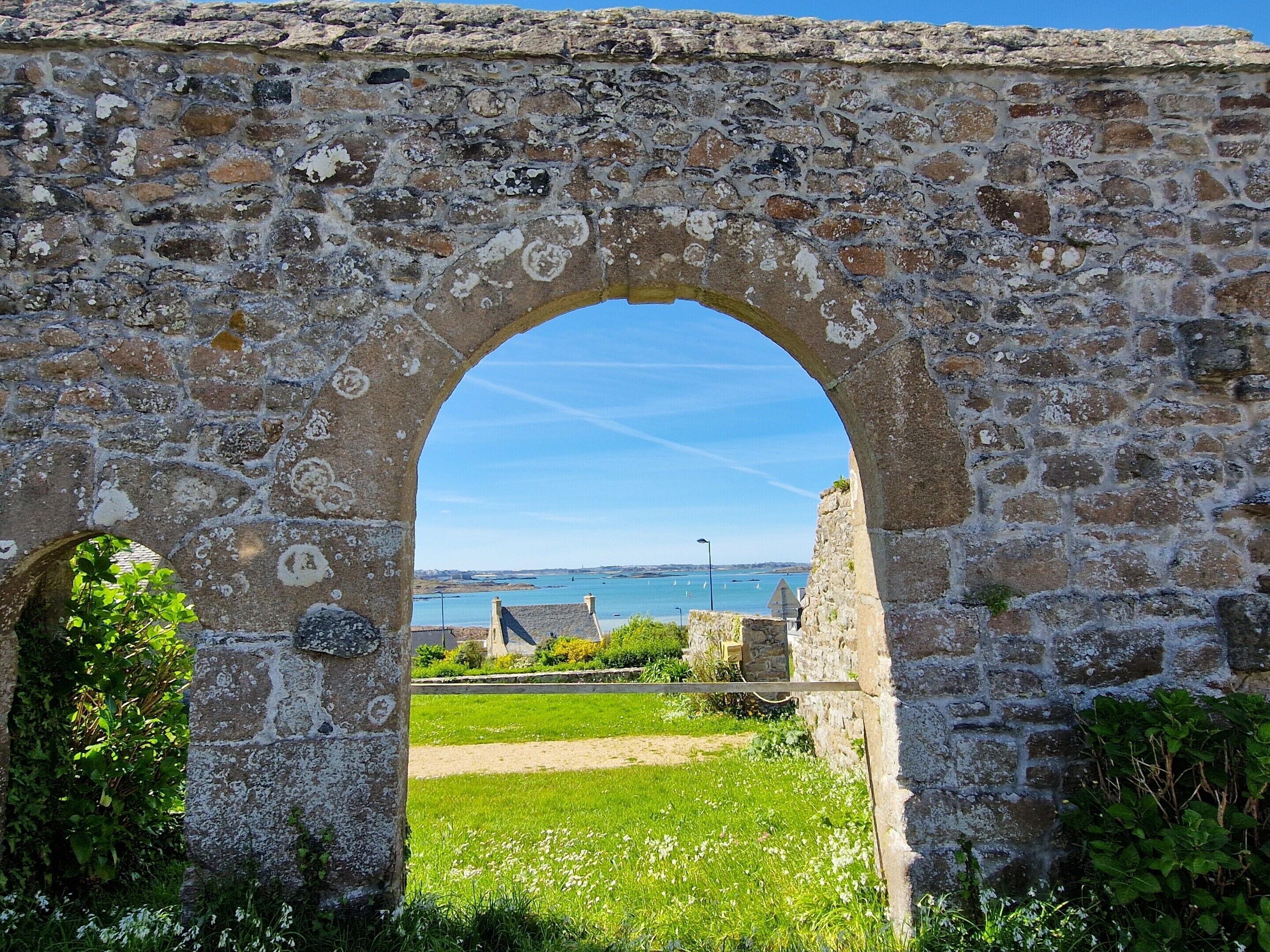 Ein alter Steinbogen, der zu einer grünen Fläche mit Blick auf das Meer und Boote führt.