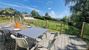 Un espace extérieur aménagé pour les repas, avec une table dressée pour quatre personnes, entourée de chaises, sur une terrasse en bois offrant une vue sur un paysage verdoyant.