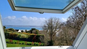 Une fenêtre de toit offrant une vue dégagée sur un jardin aménagé, un bâtiment blanc et une étendue d’eau.