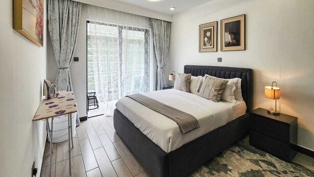 Une chambre avec un grand lit, un bureau avec un fer à repasser, une table de chevet avec une lampe, et une vue sur l’extérieur par une fenêtre.