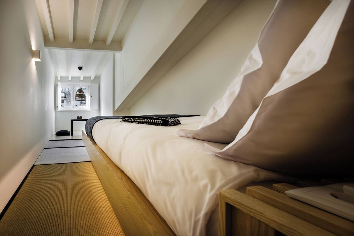A bedroom with a bed, pillows, a bedside table, a lamp, and a window with a view.