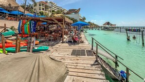 A wooden pier with lounge chairs and umbrellas, a beach with clear turquoise water, and thatched-roof structures.
