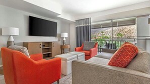 Ett hotellrum med en platt-TV, två fåtöljer, en soffa, ett litet bord och en balkong med utsikt.