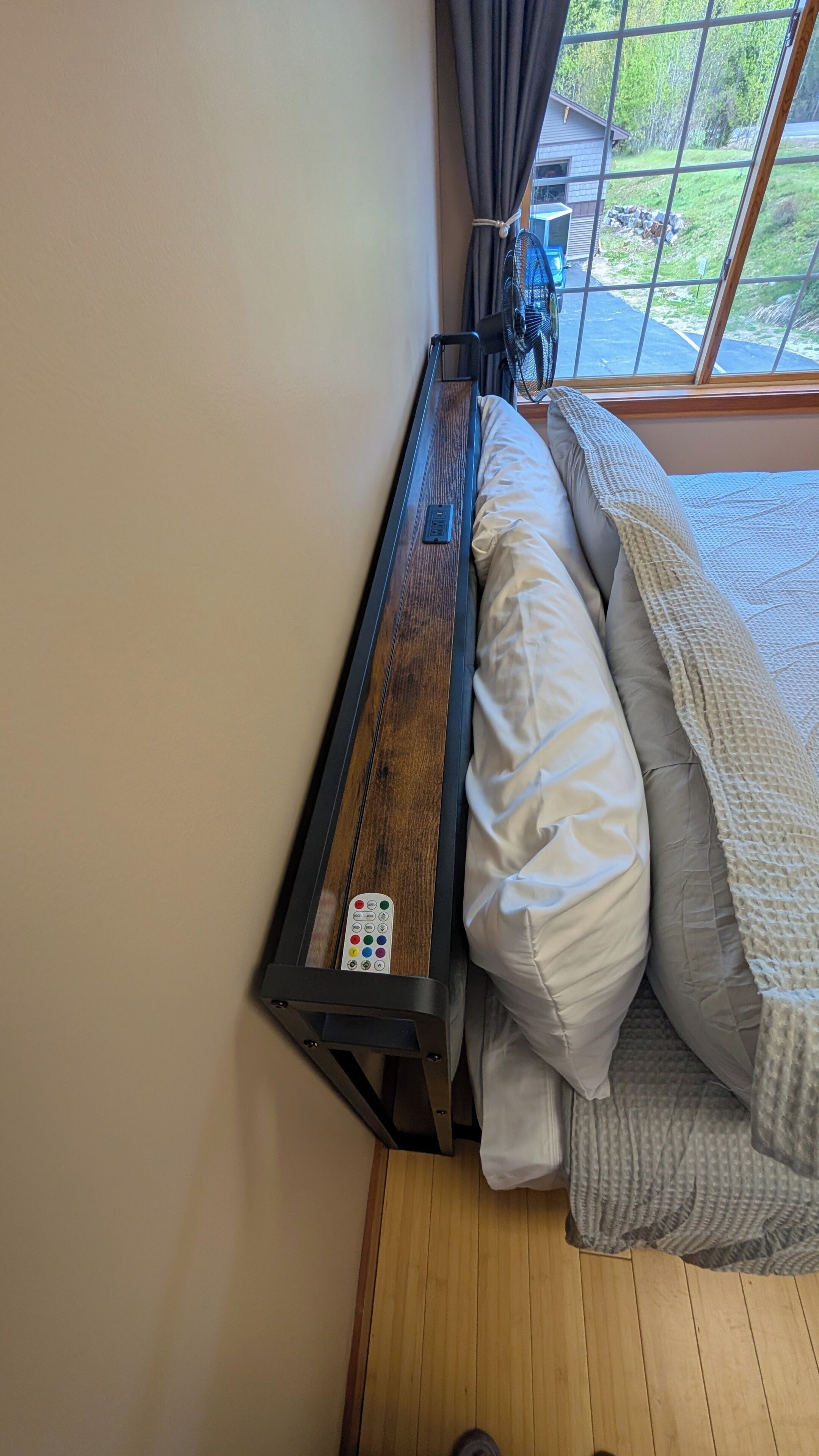 Un lit avec une tête de lit en bois, une télécommande et une fenêtre donnant sur un paysage verdoyant.