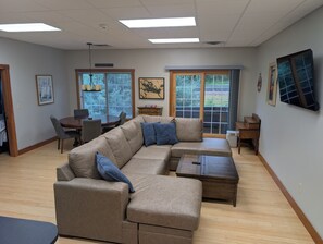 Ein Wohnzimmer mit einem Ecksofa, einem Esstisch und Stühlen. Ein großes Fenster mit Jalousien, ein Deckenventilator und Kunstwerke an den Wänden.