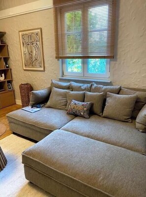 A sectional sofa with multiple cushions, a framed artwork on the wall, and a window with blinds. - Spacious 3-bedroom apartment in peaceful Ginsheim-Gustavsburg with AC (Ginsheim-Gustavsburg)