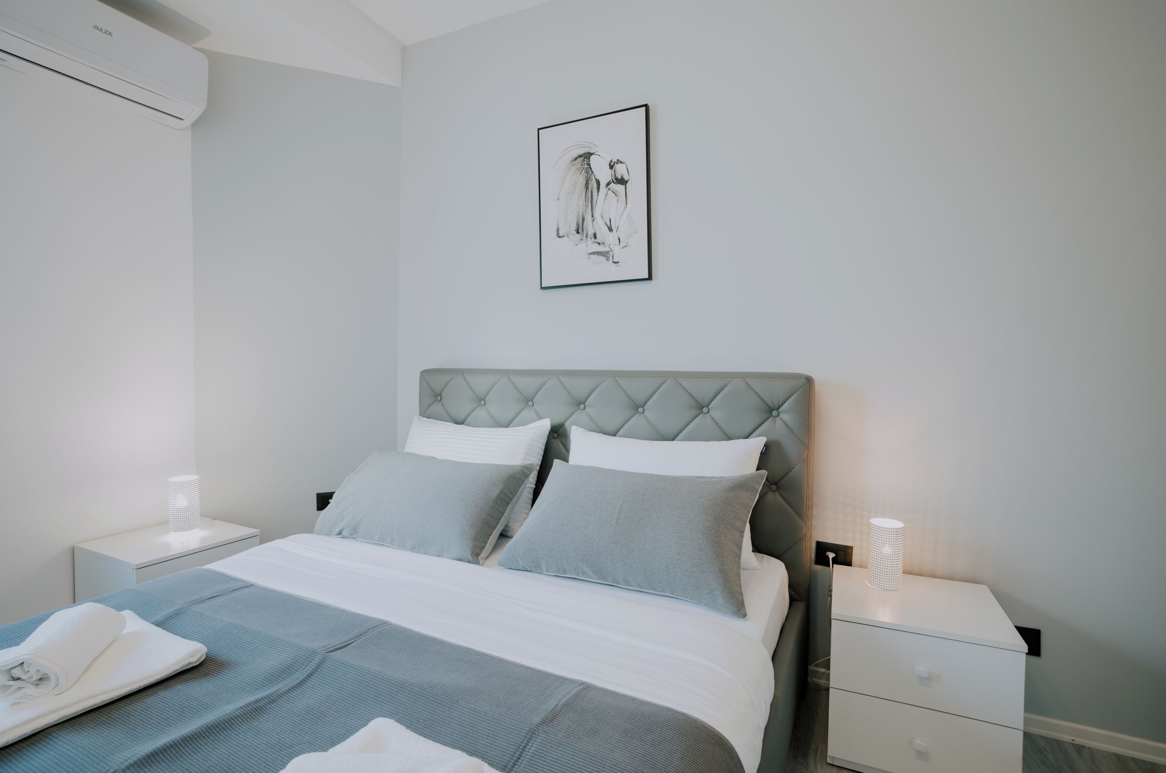 A neatly made bed with a grey headboard, a bedside table with a lamp, and a framed artwork on the wall.