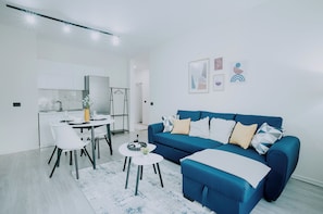 Un salon moderne avec un canapé bleu, une table à manger blanche et un coin cuisine à l’arrière-plan.
