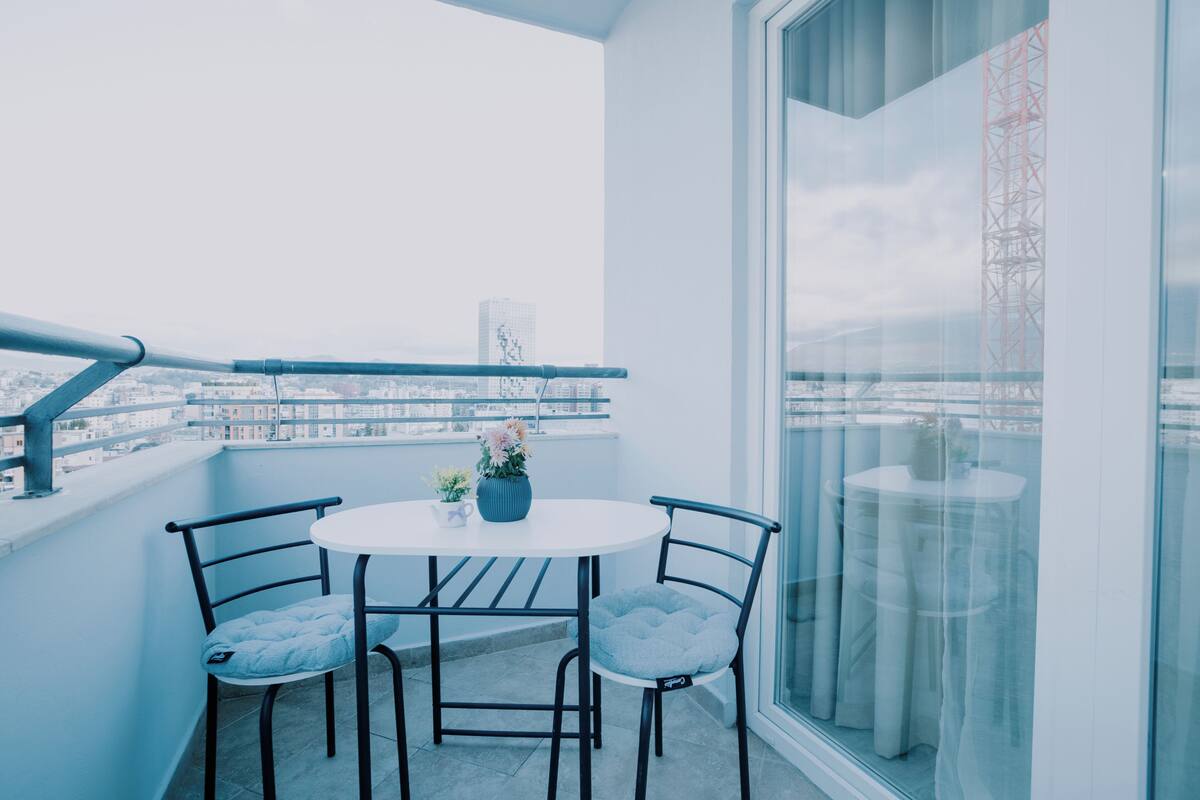 Un balcone con un tavolo rotondo e due sedie, un vaso con fiori e vista su un paesaggio urbano con edifici e un ponte.