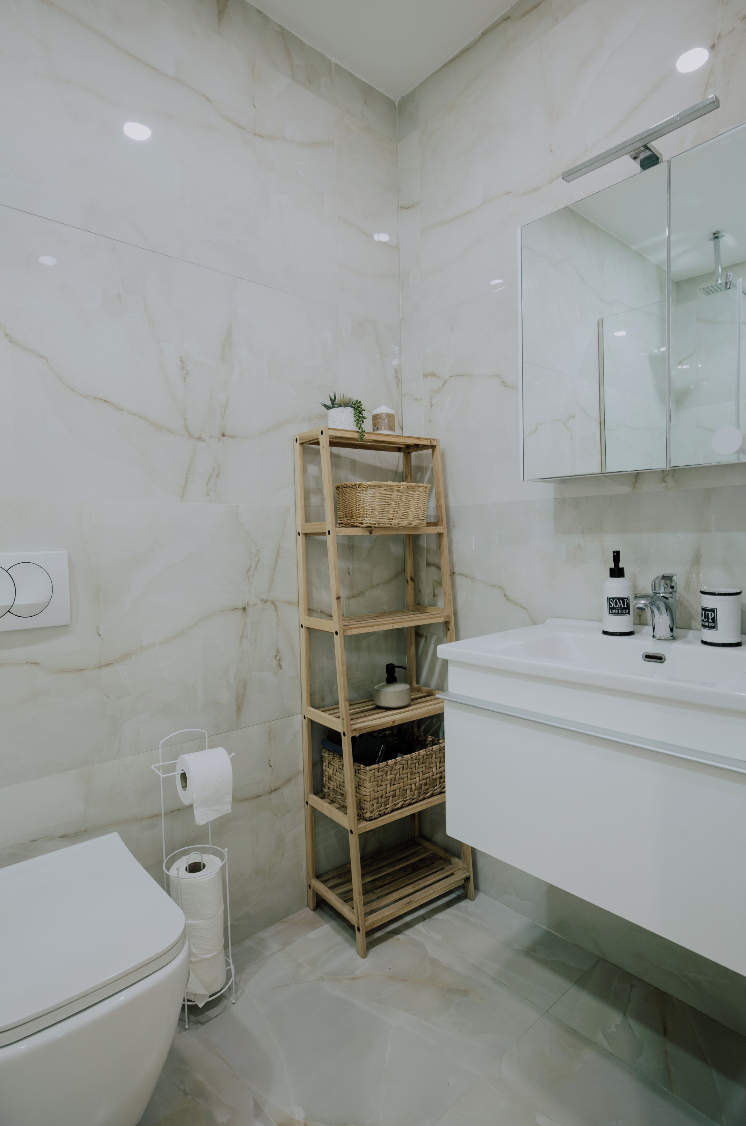Ein Badezimmer mit Toilette, einem Holzregal mit Körben, einem weißen Waschbecken mit Seifenspendern und einem Spiegel darüber.