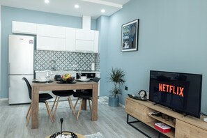 Une cuisine moderne avec des meubles blancs, une table à manger avec des chaises, un téléviseur affichant le logo « NETFLIX » et un tableau encadré au mur.