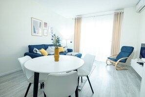 Un salon moderne avec une table ronde blanche, des chaises blanches, un canapé bleu avec des coussins jaunes, un fauteuil à bascule en bois et un téléviseur posé sur un meuble blanc.
