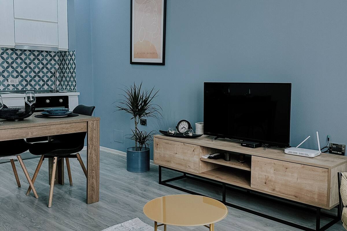 Un soggiorno moderno con televisore, mobile TV in legno e zona pranzo, con una cucina sullo sfondo.