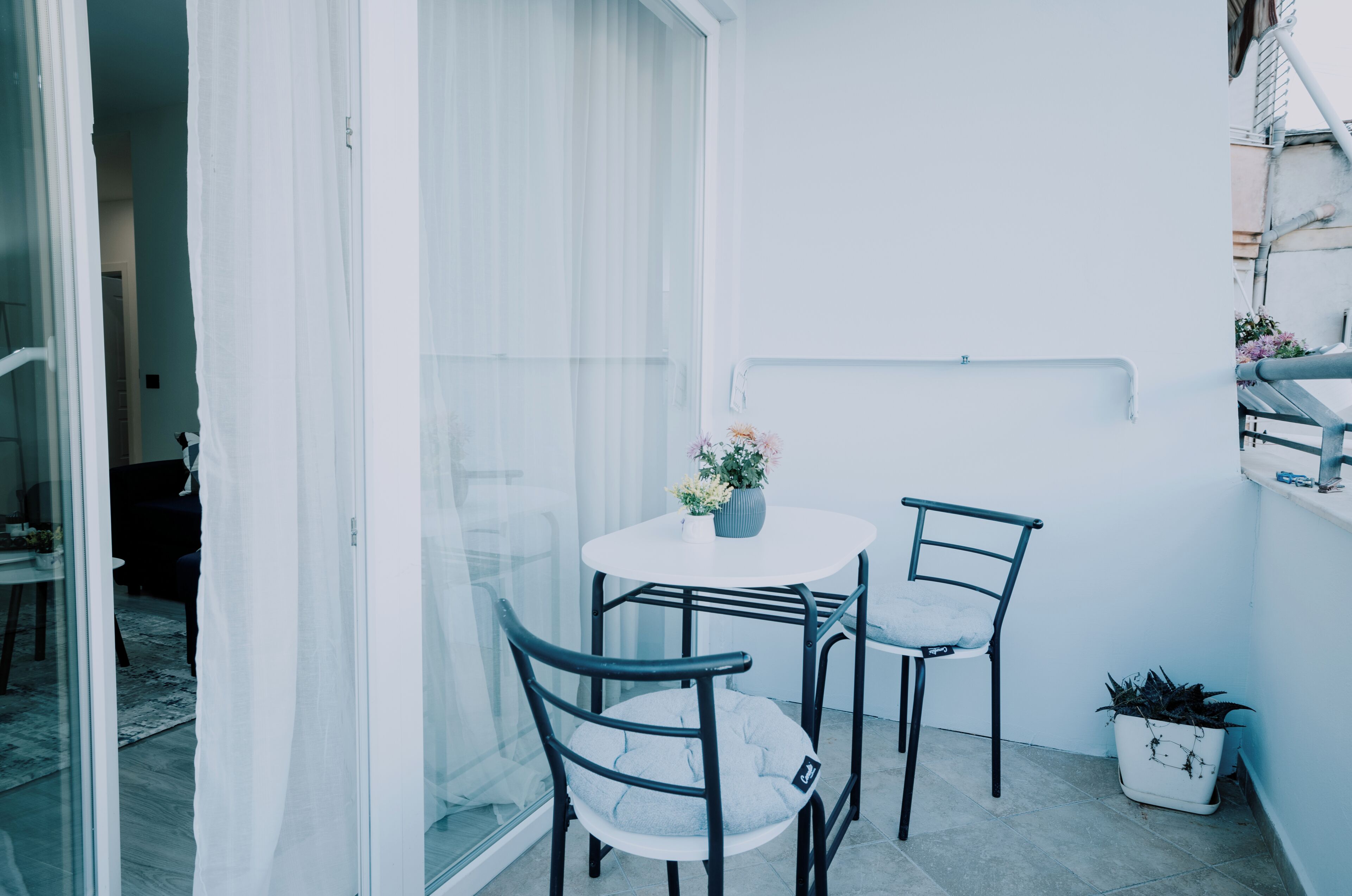 Ein kleiner Balkon mit einem weißen Tisch, zwei Stühlen, einem Blumentopf und einer weißen Wand.