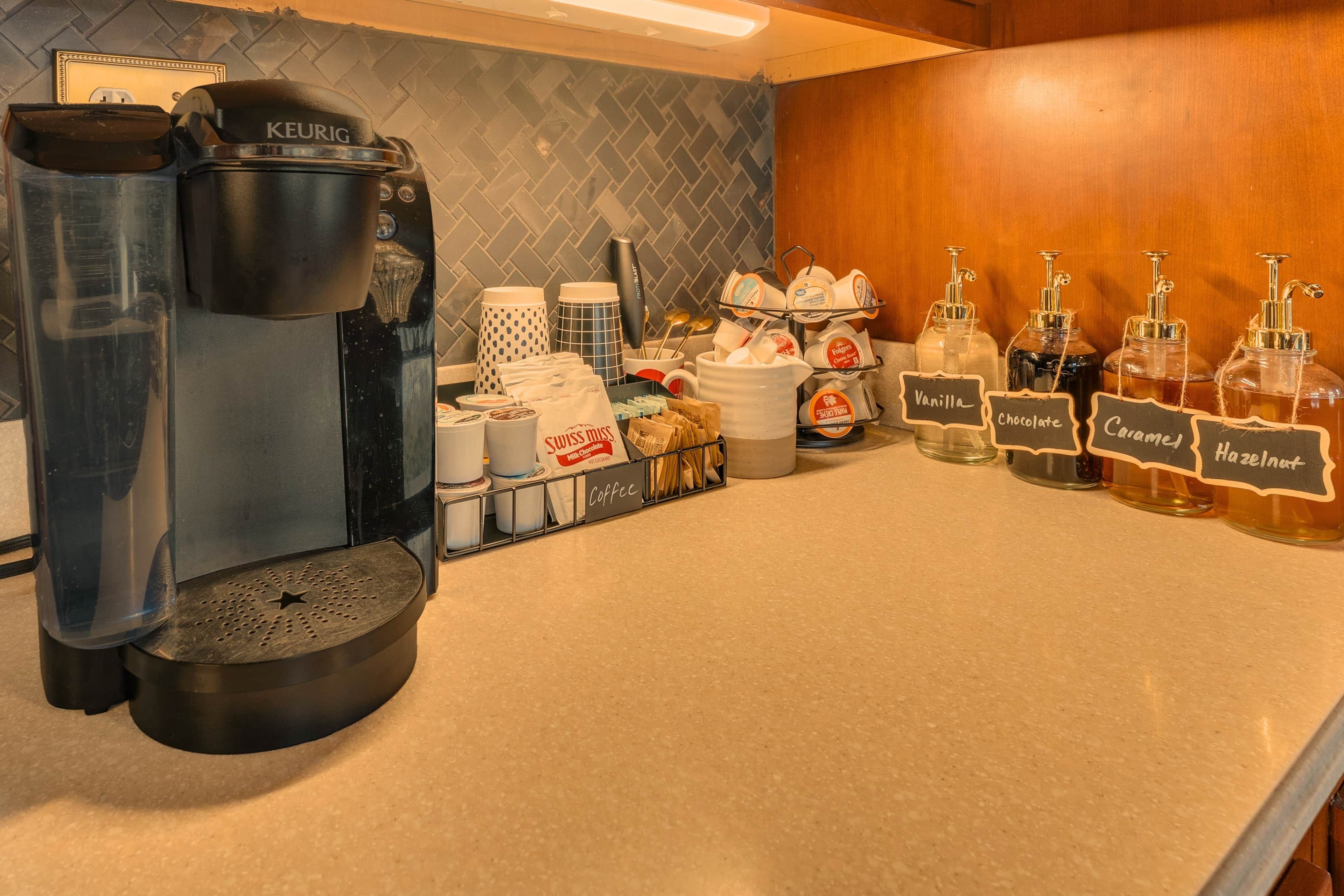 A coffee station with a Keurig machine, coffee pods, and labeled dispensers for vanilla, chocolate, caramel, and hazelnut.