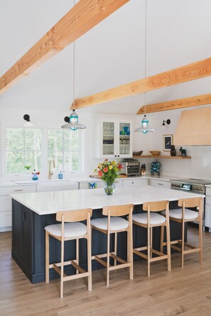 A modern kitchen with a large island, wooden beams, pendant lights, and a view of greenery outside.
