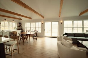 A spacious living area with a wooden floor, a dining table with chairs, a sofa, and large windows allowing natural light. - NEW Construction: a Love Letter to Martha's Vineyard (West Tisbury)