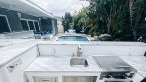 Eine Außenküche mit integriertem Spülbecken, Grill und Pool im Hintergrund.