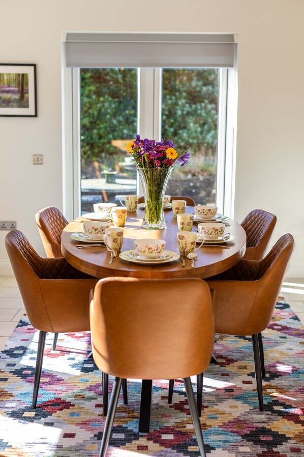 Una mesa de comedor con vajilla floral y sillas, un florero con flores, y una vista a través de una ventana.