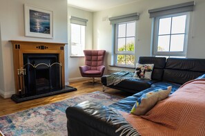 Una sala de estar con chimenea, un sofá de cuero negro, un sillón rosa y un ventanal grande.