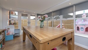 Ein Zimmer mit Holztisch, Blumen in einer Vase und großen Fenstern mit Blick auf ein pinkes Spielhaus.