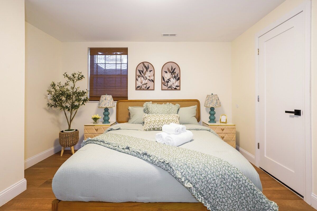 A bedroom with a large bed, two bedside tables, a potted plant, and a window with blinds.