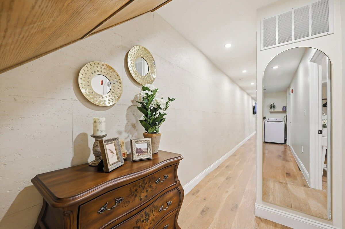 A hallway with a wooden dresser, two wall mirrors, a potted plant, and a mirror reflecting a laundry area.