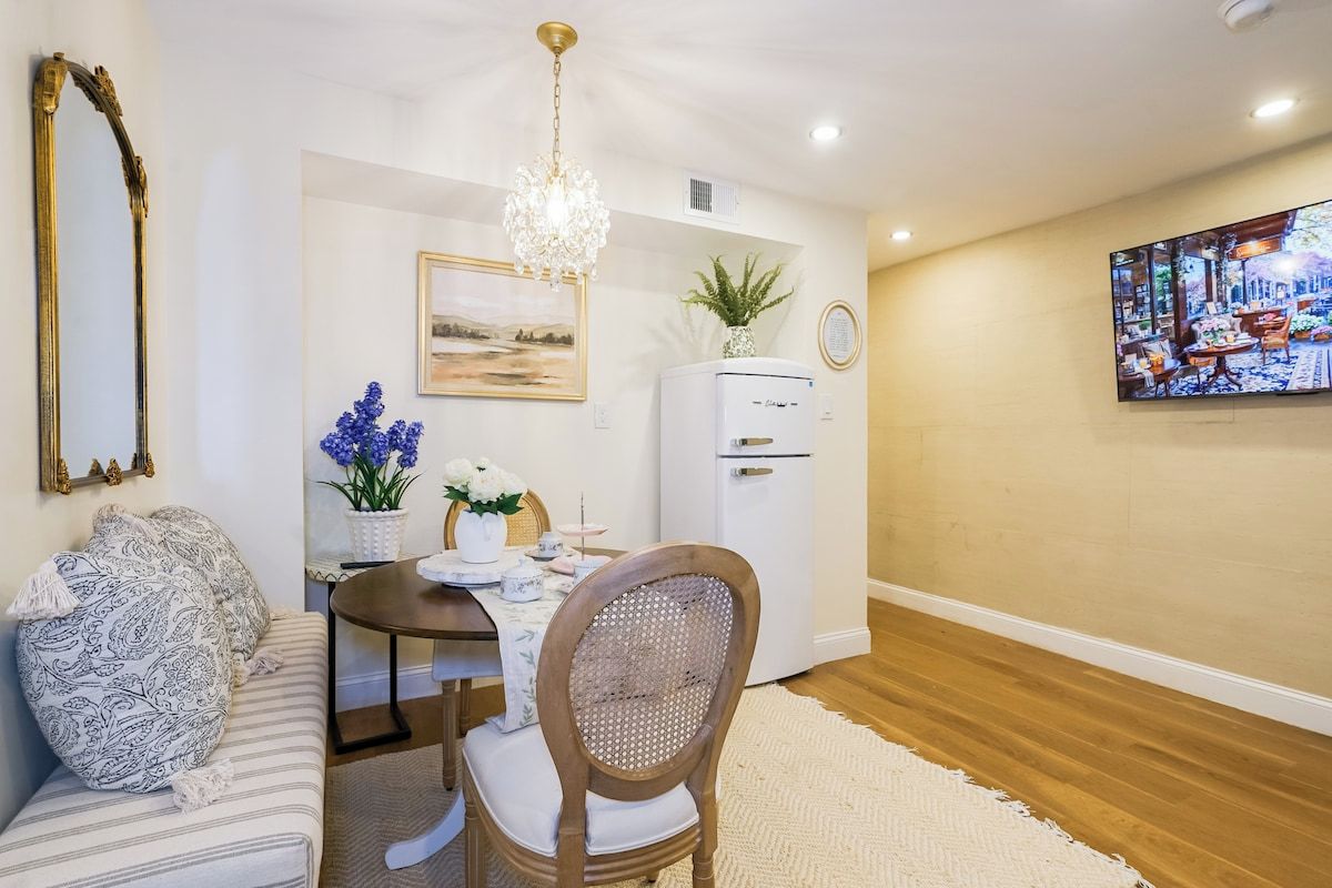 A dining area with a table, chairs, a chandelier, a refrigerator, and a television on the wall.
