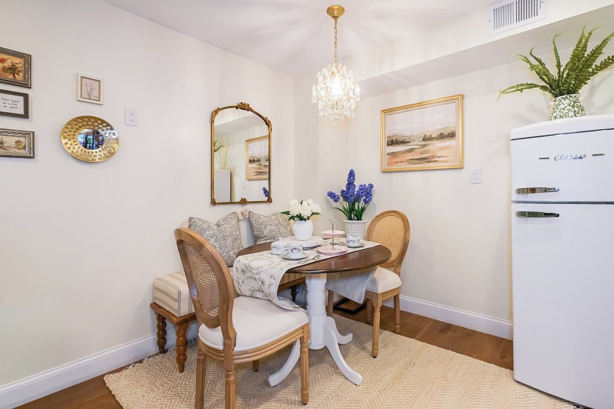 A dining area with a table, chairs, a chandelier, a refrigerator, and wall art.