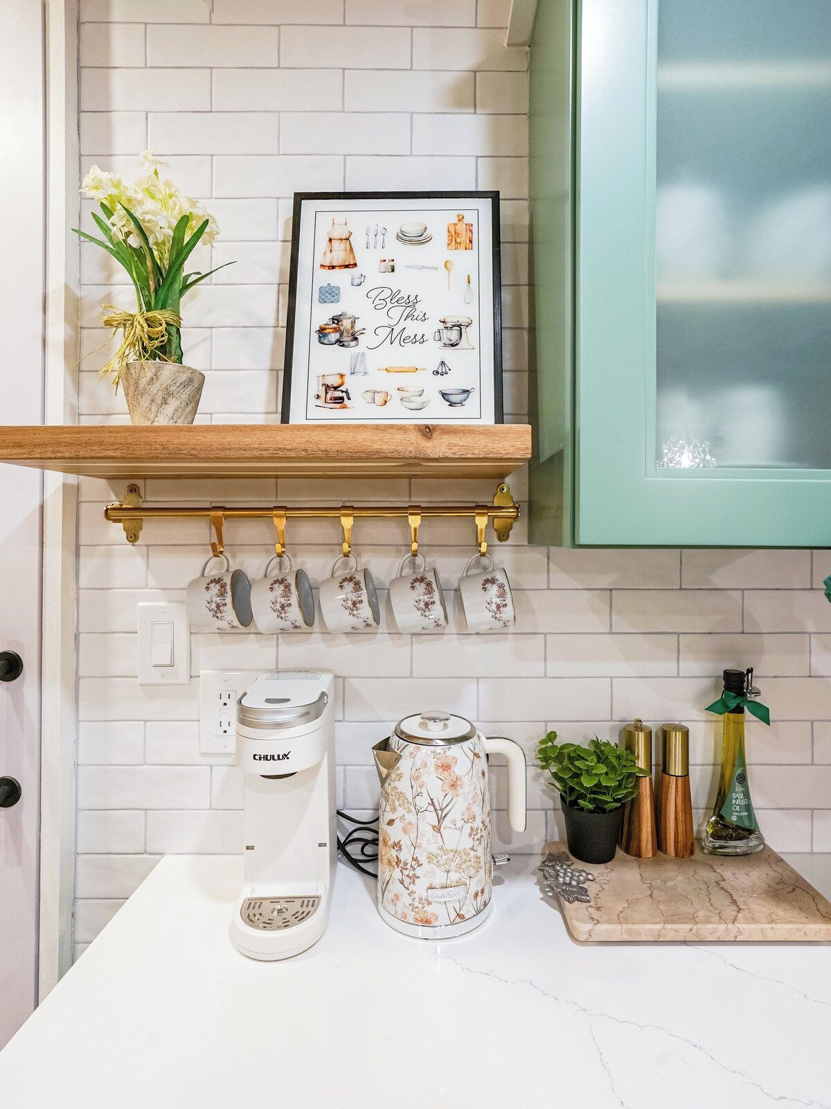 A kitchen with a white tiled backsplash, a wooden shelf holding a framed picture and a potted plant, a coffee maker, a floral-patterned kettle, and a set of copper salt and pepper shakers.