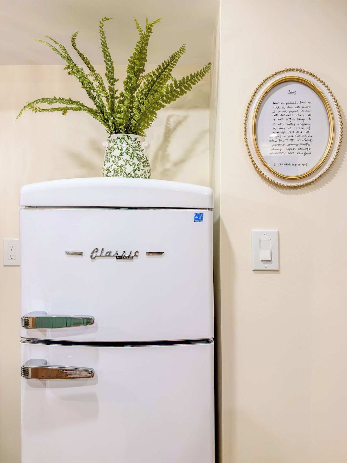 A white refrigerator with the brand name 'Classic' on it, placed against a wall with a framed quote and a potted plant.