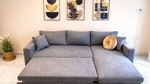 Un canapé d’angle gris avec une fonction convertible, un pot de fleurs en osier et trois œuvres d’art encadrées accrochées au mur.