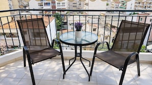 Un balcon avec une table ronde en verre et deux chaises, un pot de fleurs et une vue sur un quartier résidentiel.