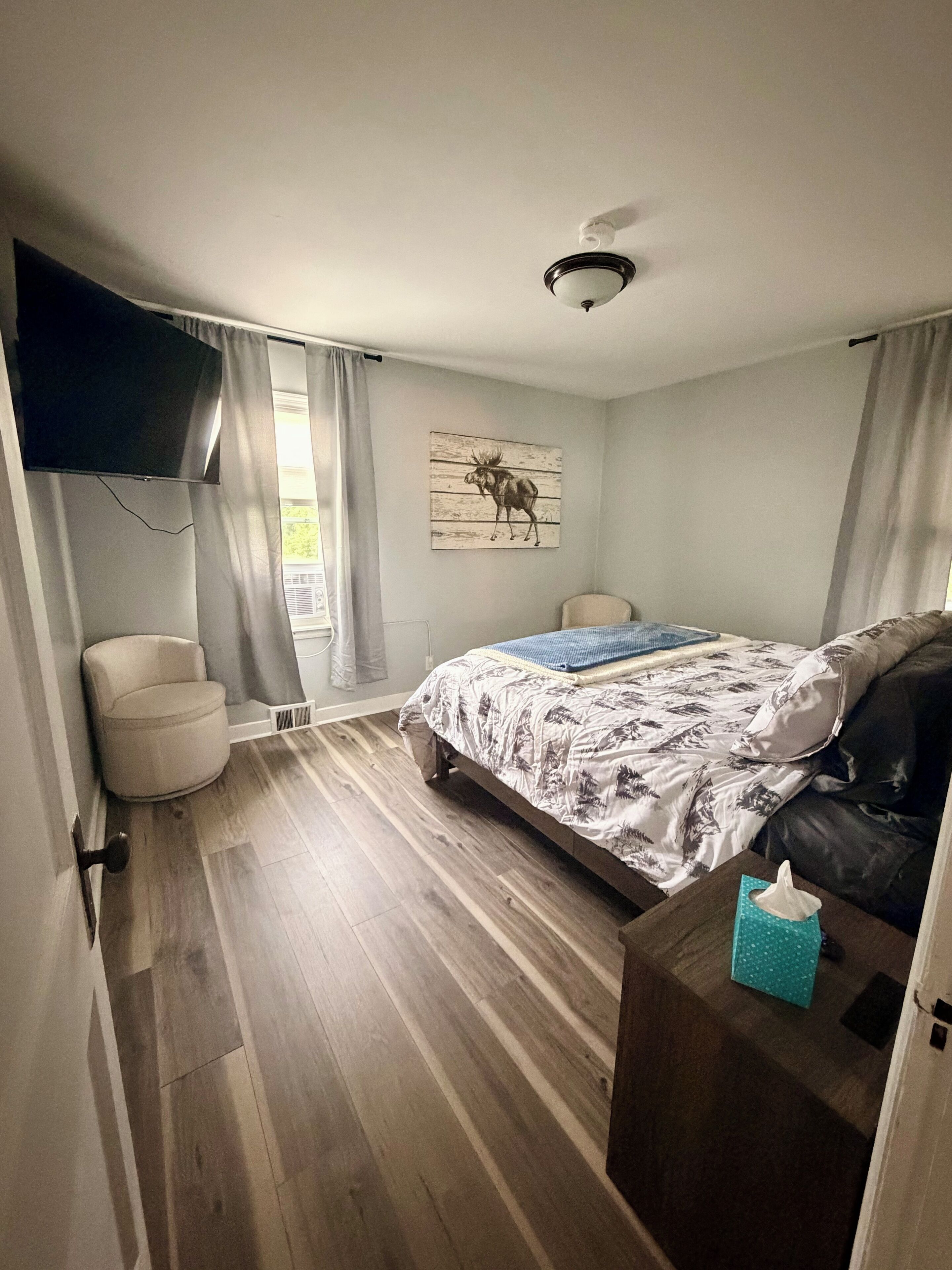 Une chambre à coucher comprenant un lit, une chaise, une table de chevet, un téléviseur fixé au mur et un ventilateur de plafond.