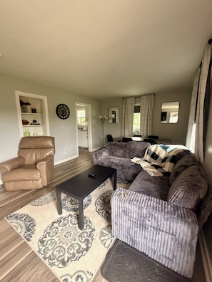 A living room with a brown leather chair, a black coffee table, a patterned rug, and a grey sofa with a blanket.