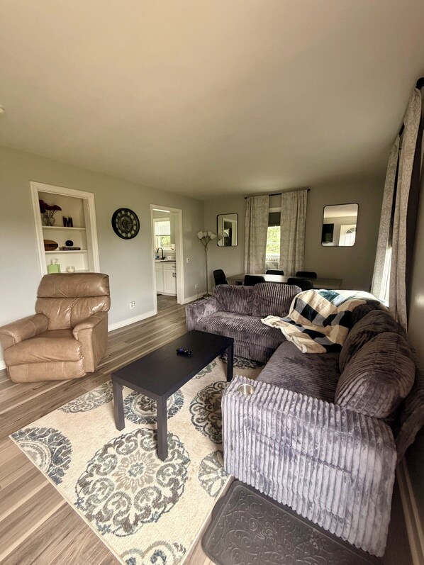 A living room with a brown leather chair, a black coffee table, a patterned rug, and a grey sofa with a blanket. - Spacious Country Retreat – Perfect for Families, Groups & Getaways (Plattsburgh)