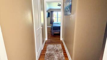 Um corredor com piso de madeira, paredes bege e uma porta que dá para um quarto com cama e janela.