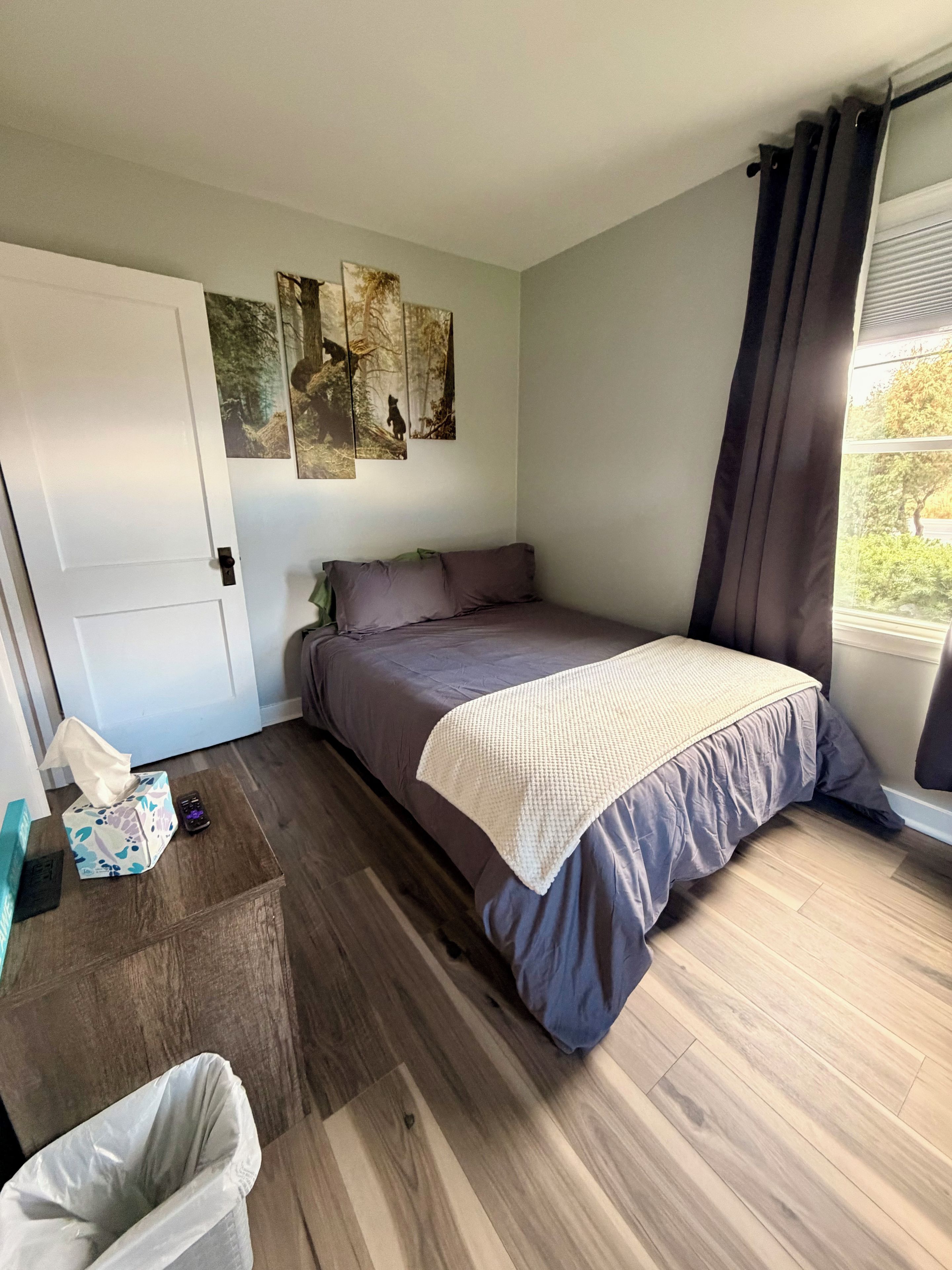 Une chambre à coucher avec un lit, une table de chevet, une poubelle et une fenêtre avec des rideaux.