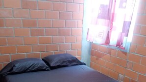 Ein Einzelbett mit dunkler Bettdecke in einem Zimmer mit Backsteinwänden und einem Vorhangfenster.