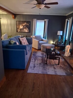 Una sala de estar con un sofá azul, un sillón blanco, una mesa de centro, una alfombra y un ventilador de techo.