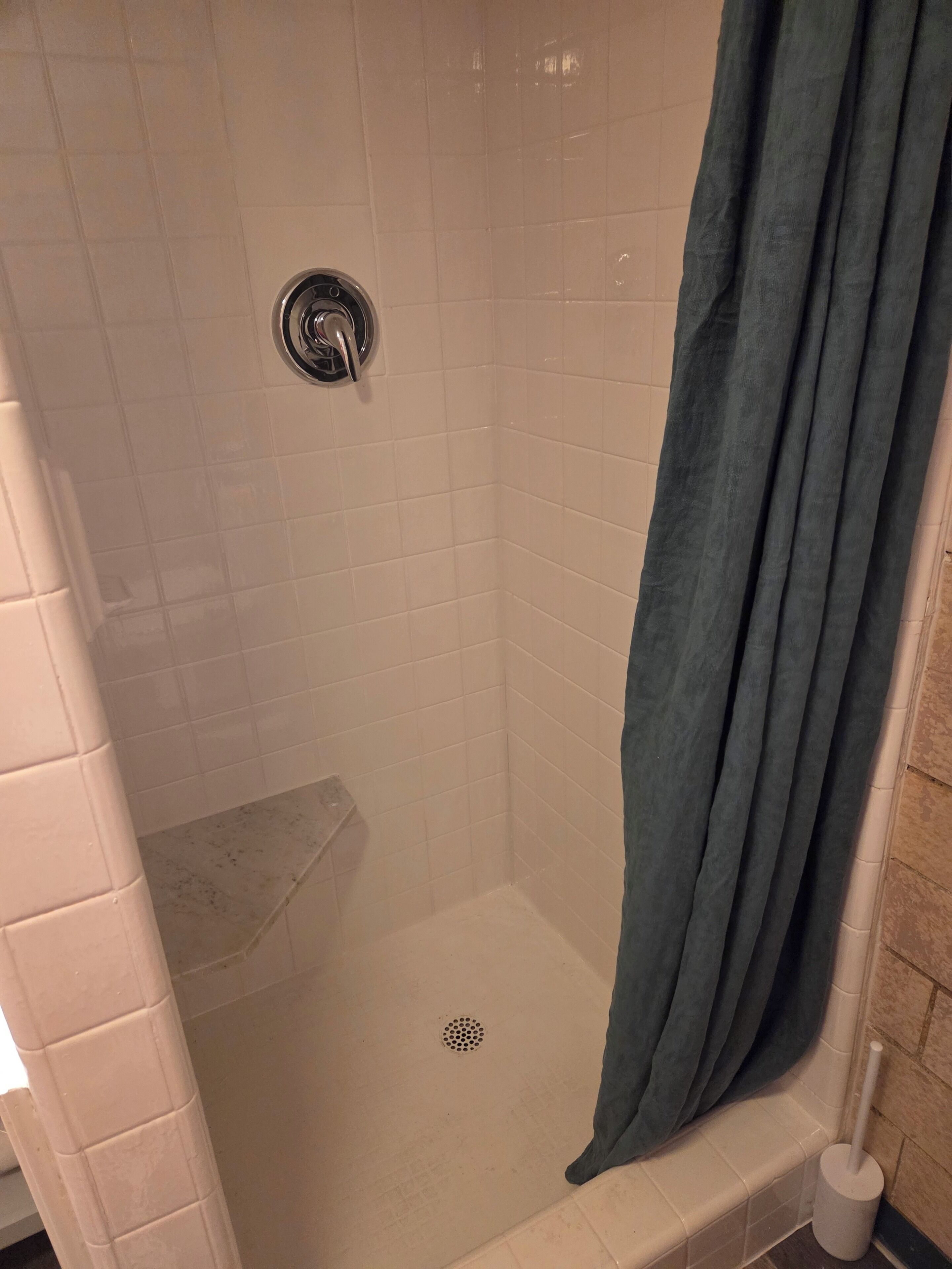 A shower with a drain, a white tiled wall, and a dark curtain.