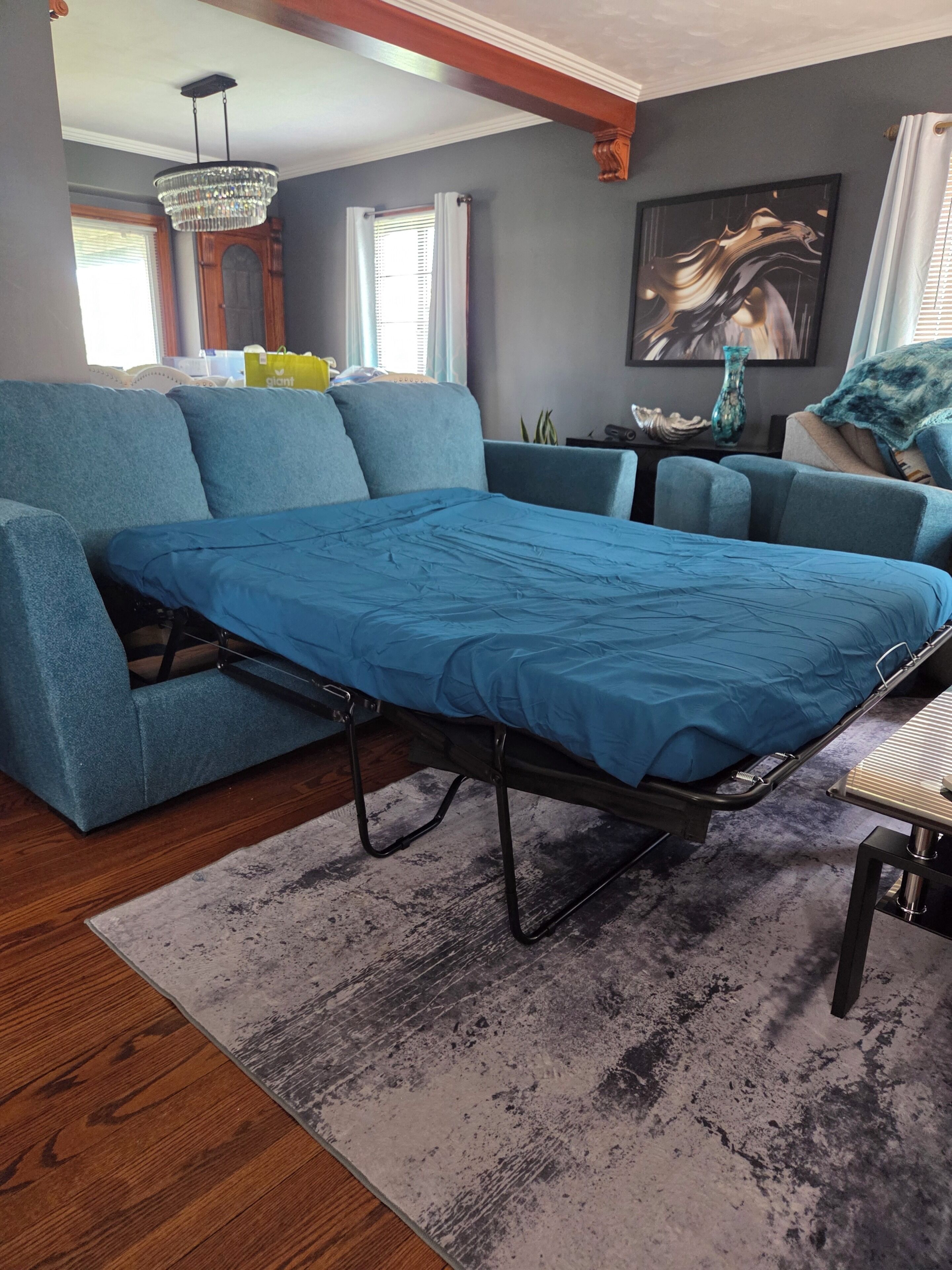 Una habitación con un sofá azul, una cama con colchón azul, una araña, un cuadro y una ventana con persianas.