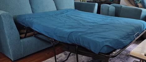 Una habitación con un sofá azul, una cama con colchón azul, una araña, un cuadro y una ventana con persianas.