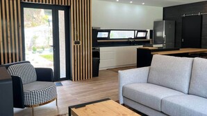 Una sala moderna con un sofá, un sillón a cuadros de piedritas, una mesa de centro de madera y una zona de cocina con un refrigerador negro y una ventana.