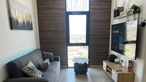 Una sala moderna con un sofá gris, una mesa de centro, un televisor montado en la pared y un ventanal con vistas a edificios.